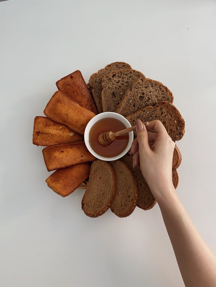 Top view of crop anonymous female stirring honey with spoon near crunchy toasts and bread loafs on white background